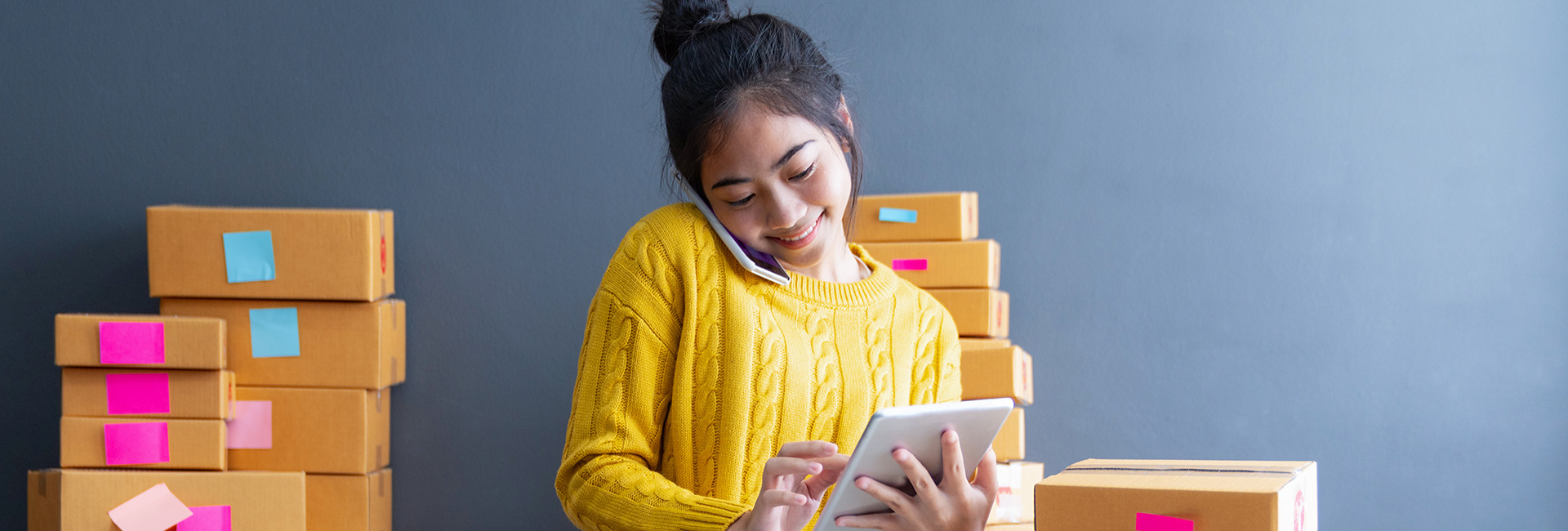 South Asian female in yellow jumper smiling with stacked boxes and tablet