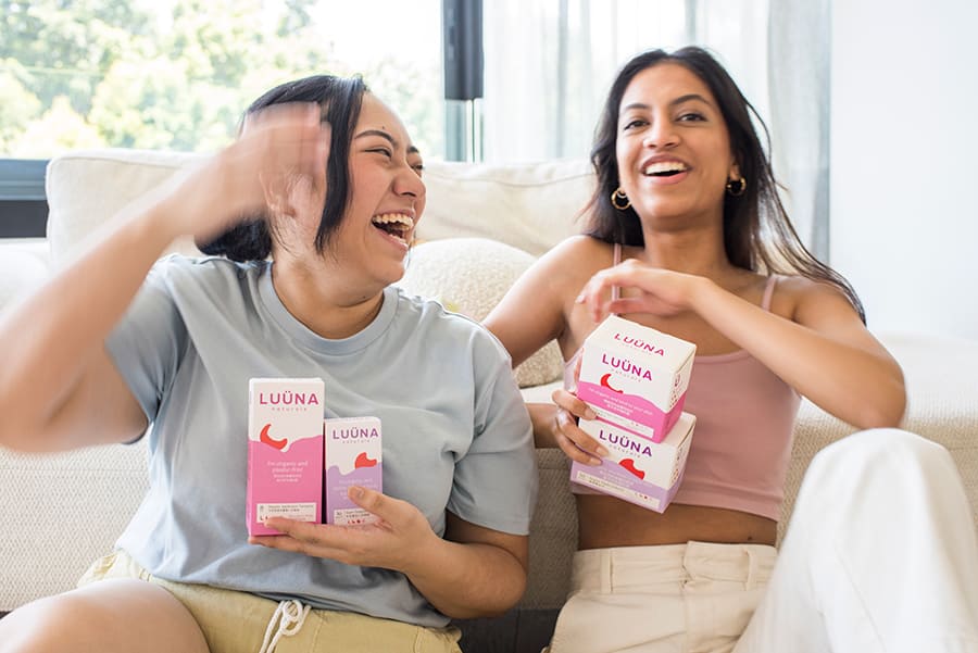 2 young Asian females sit on sofa laughing holding period products