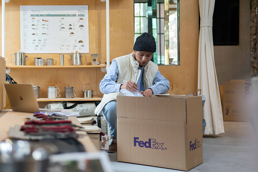 Young Asian male in woollen hat packing FedEx boxes in shop