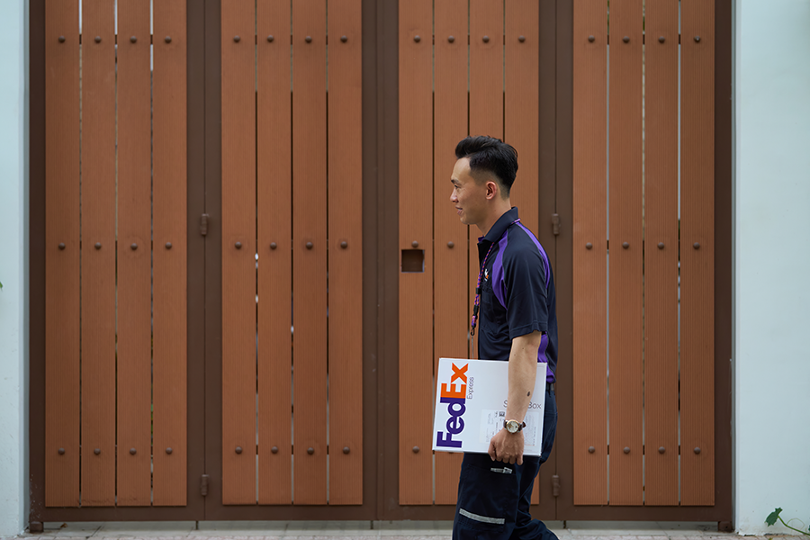 Male Asian FedEx courier carries parcel outside brown gates