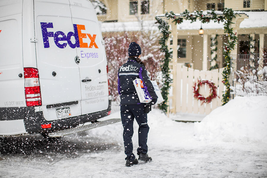 Female FedEx courier delivers parcel to white house with festive wreath in snow