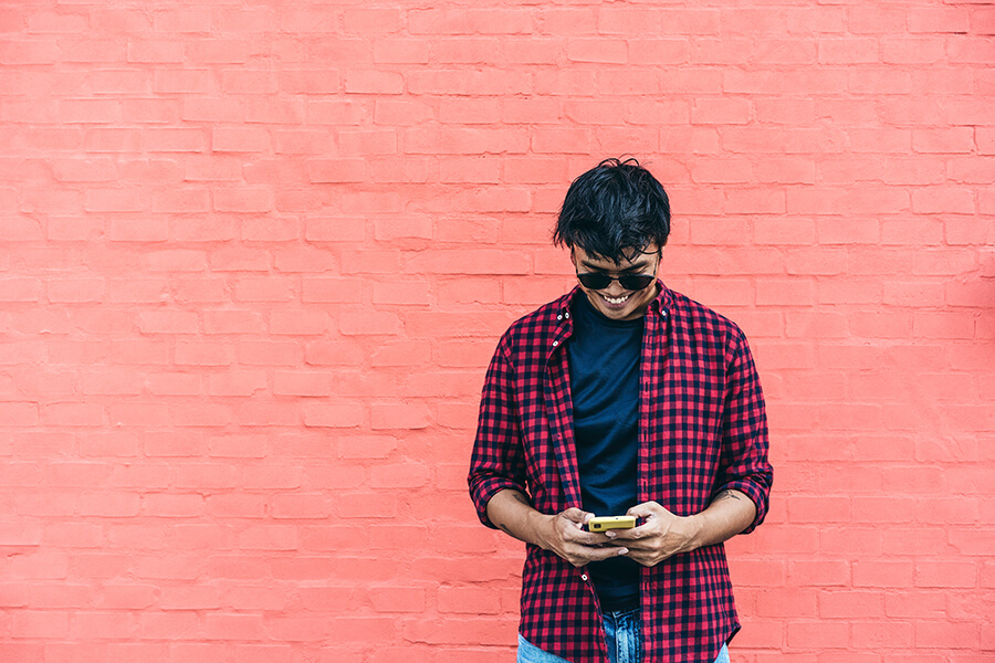 Asian male in red checked shirt smiles at phone in front of pink wall