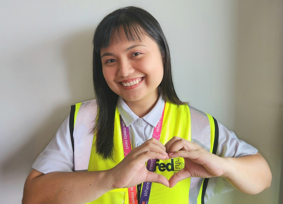 Smiling Asian female in high-vis uniform stands in front of FedEx sign making heart gesture