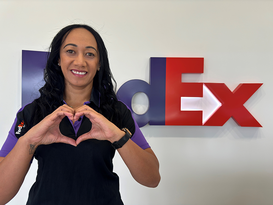 Smiling dark-haired female stands in front of FedEx sign making heart gesture