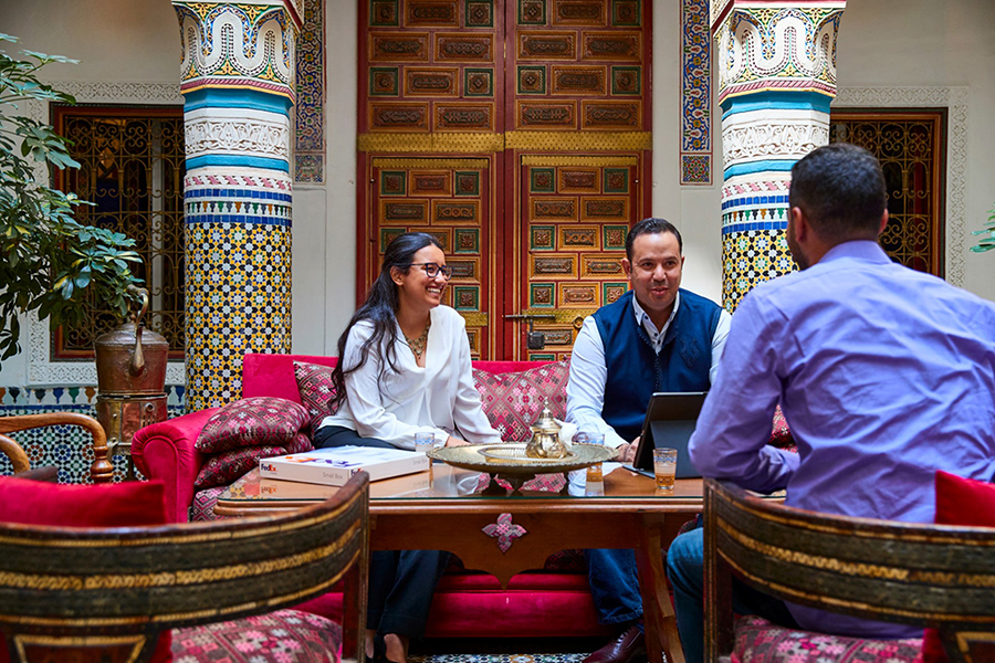 Business meeting in a traditional Arab setting with colorful mosaic and intricate woodwork, featuring two men and a woman discussing over a table, exuding a blend of cultural heritage and modern commerce.