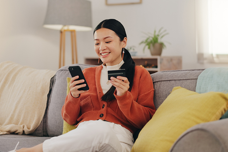 Young Asian female sits on sofa with mobile phone and credit card
