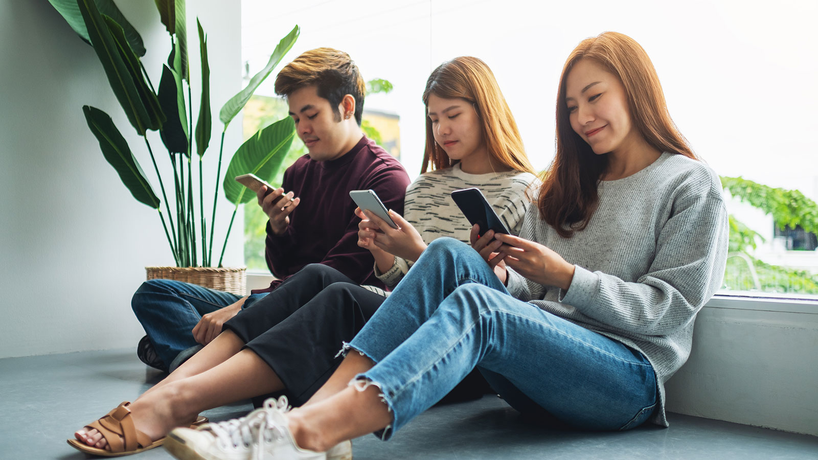 A group of millennials, a male and two Asian females, sit leaning against wall looking at phone