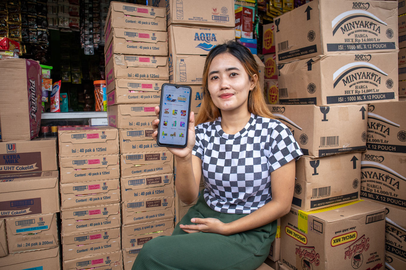 Indonesian female shopkeeper surrounded by boxed stock holding up phone