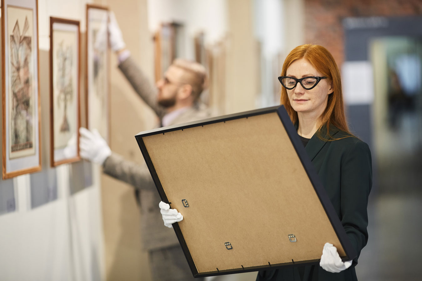 Red-headed female exhibition curator wearing white gloves hangs artwork in gallery