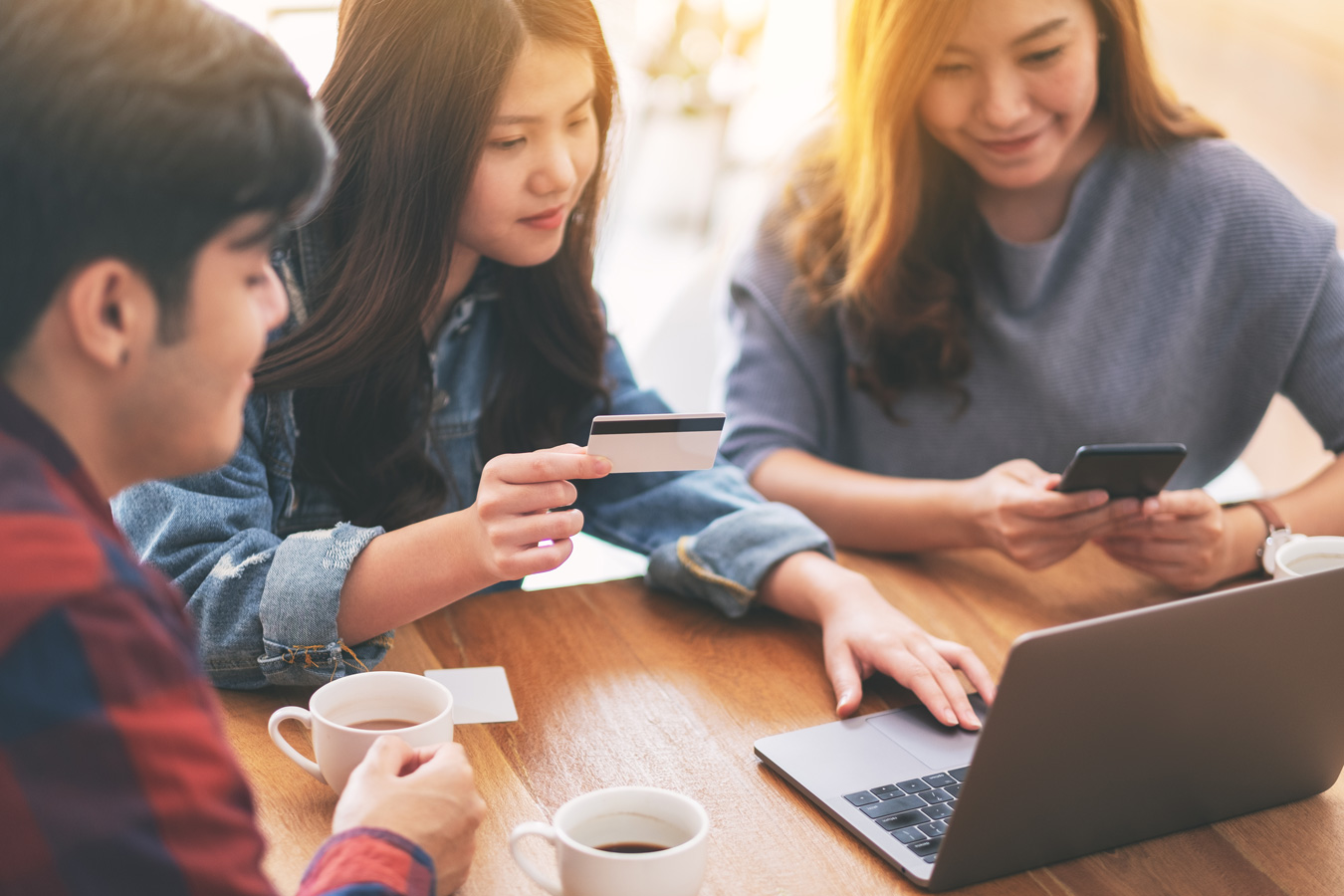 Three young Asian friends using credit cards to make online laptop purchase