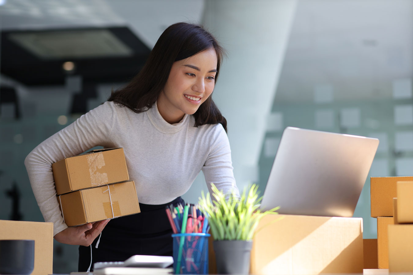 Young Asian female business owner holding small packages and using laptop