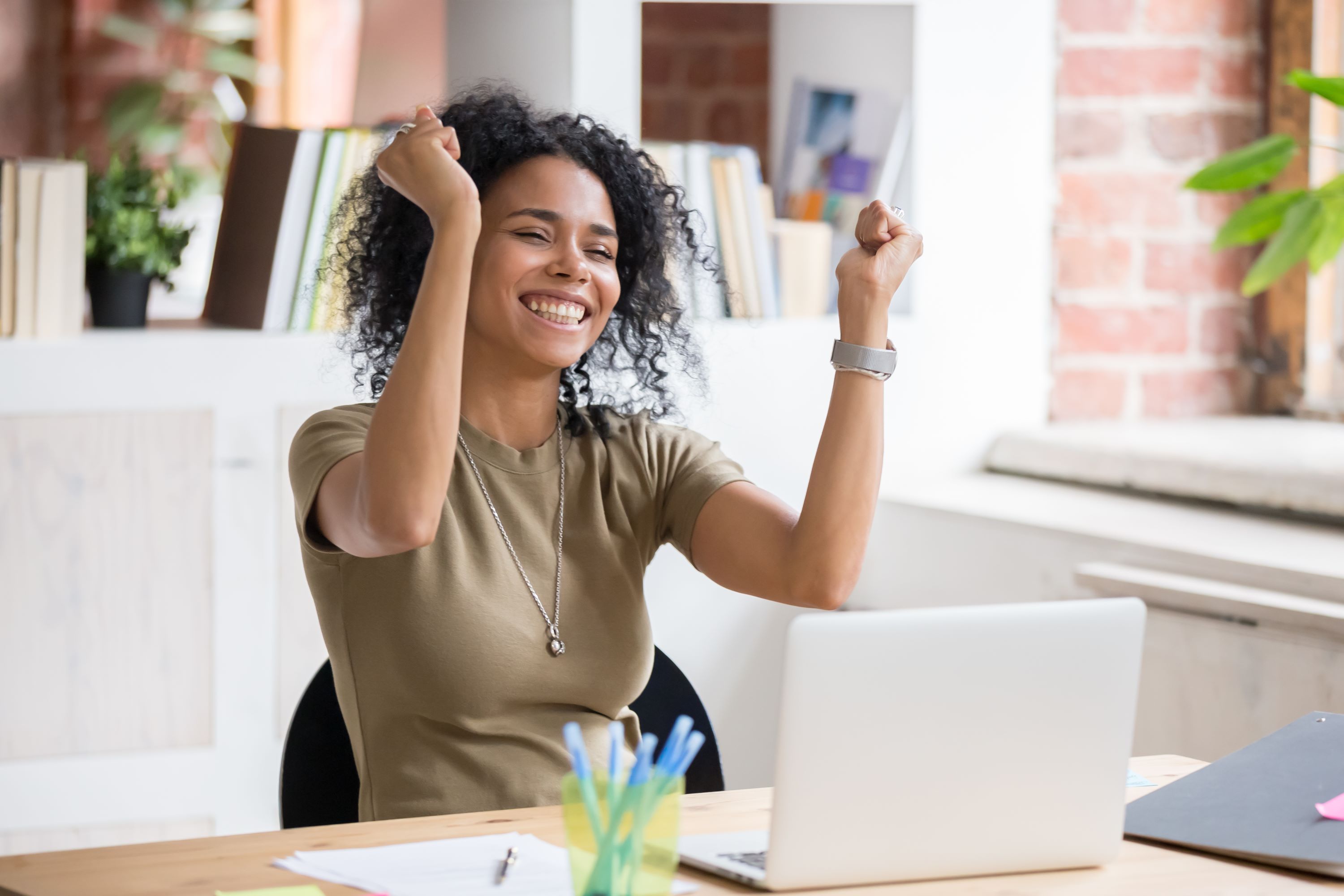 Young female with dark curly hair celebrates by raising both hands at her work desk